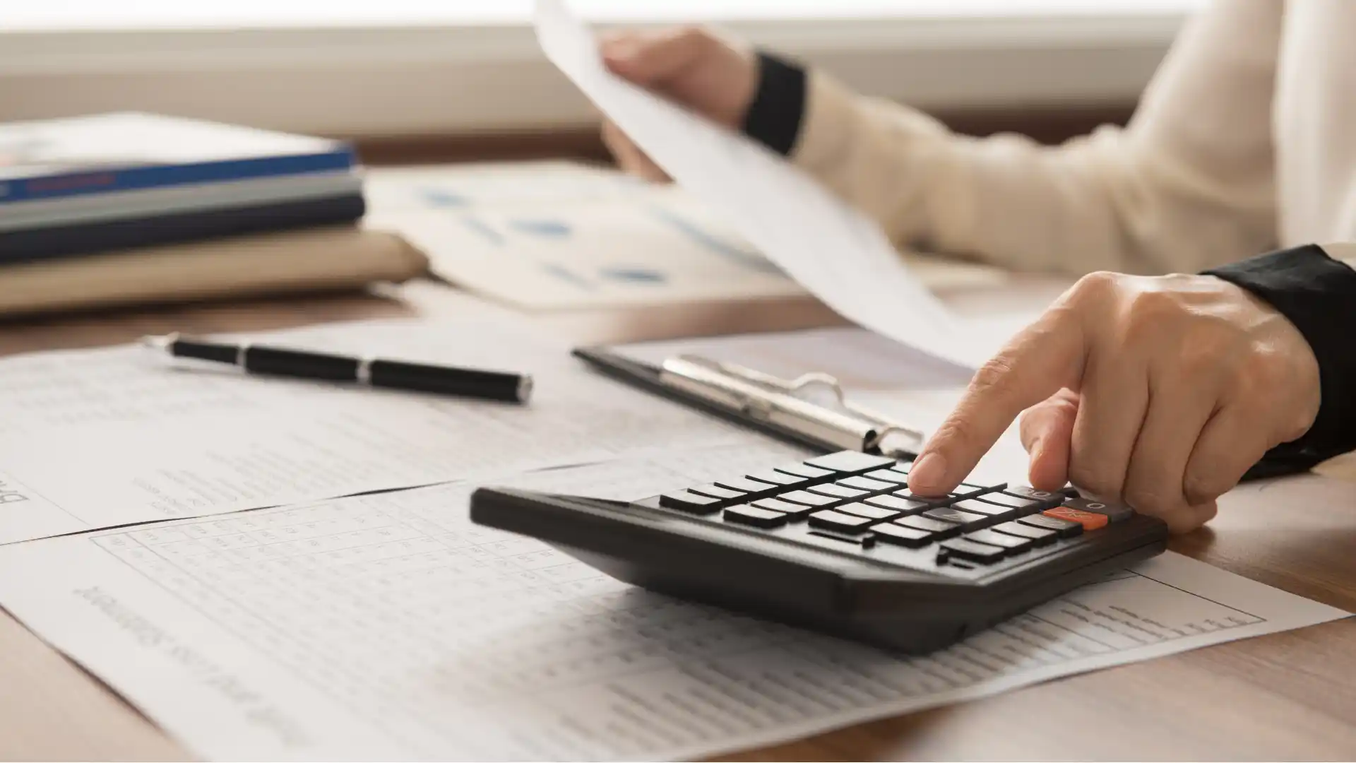 Hand using a calculator to review financial statements and tax reports on a desk.