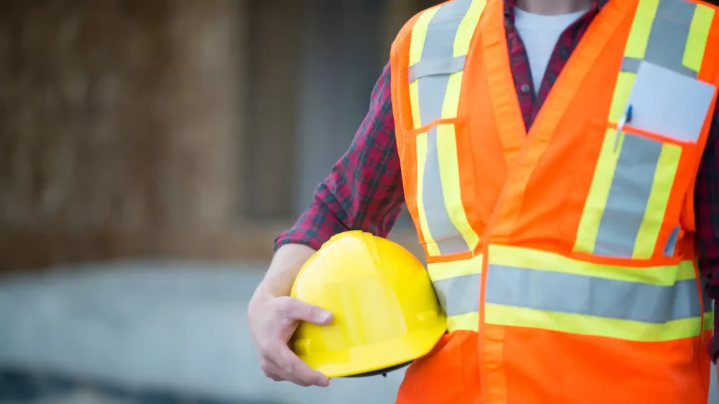 Construction worker wearing a high-visibility vest holding a yellow hard hat at an industrial job site.
