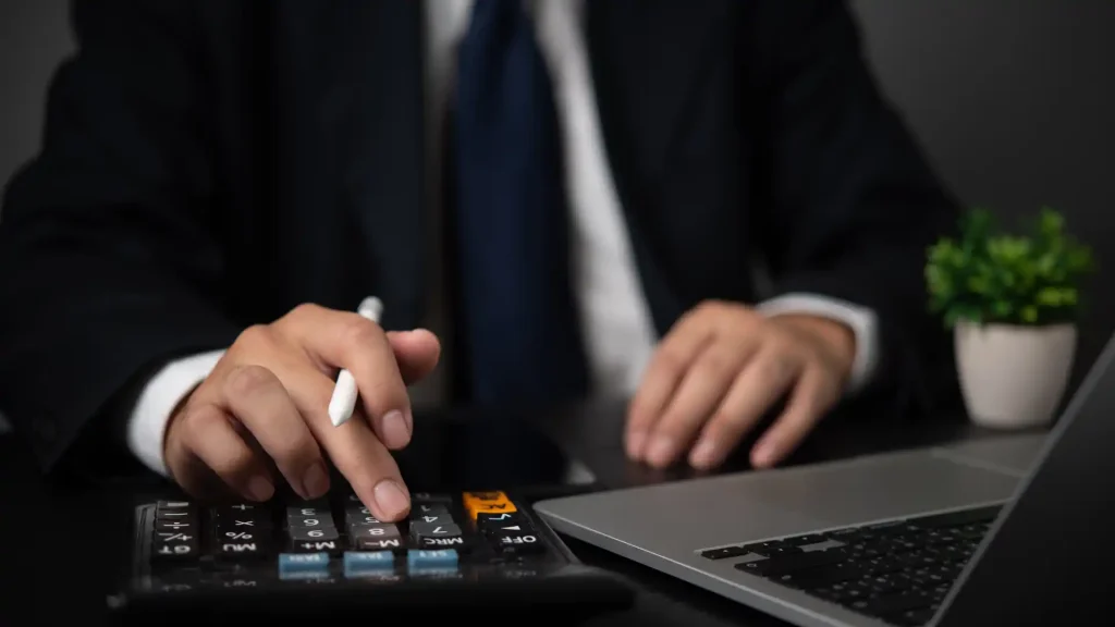 Business professional calculating financial data on a desk with a calculator, laptop, and plant, representing back office and tax compliance services in Mexico.