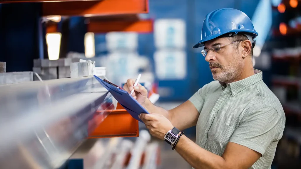Industrial supervisor wearing a blue hard hat inspecting materials and taking notes in a manufacturing plant in Mexico.