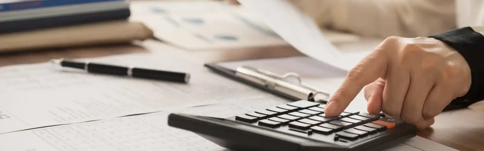 Hand using a calculator to review financial statements and tax reports on a desk.
