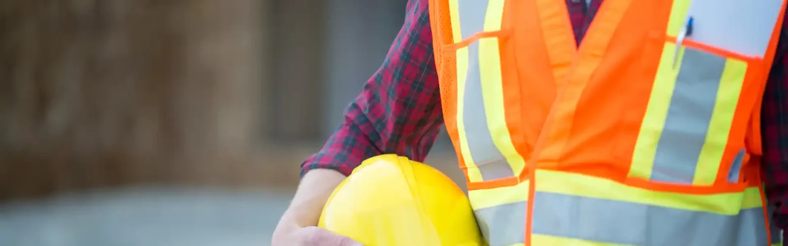 Construction worker wearing a high-visibility vest holding a yellow hard hat at an industrial job site.