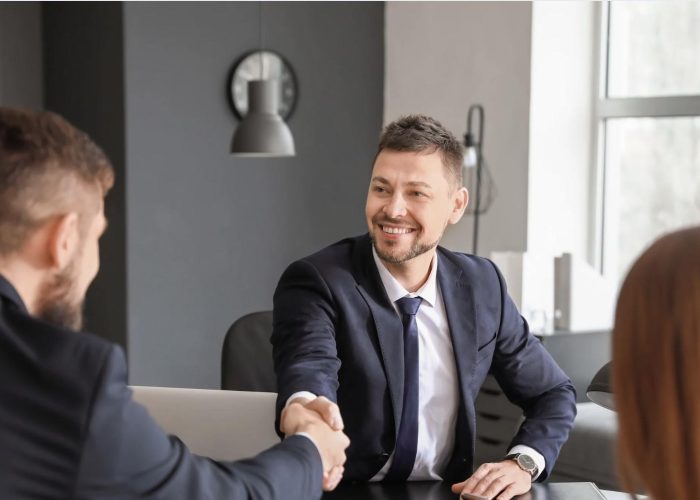 Two businessmen in suits shaking hands across a desk, smiling, with a woman present in the meeting, symbolizing successful business negotiations and partnerships in Mexico.
