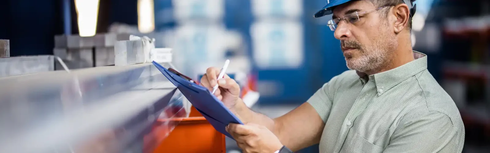 Quality Control and Operational Supervision in Industrial Operations in Mexico Industrial supervisor wearing a blue hard hat inspecting materials and taking notes in a manufacturing plant in Mexico.